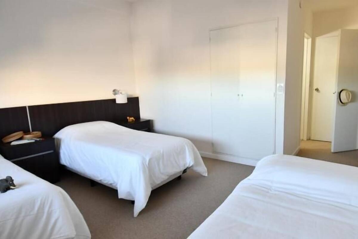 A hotel room with two single beds, a dark headboard, a bedside table with a lamp, and a door leading to another room.