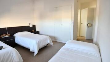 Quarto de hotel com duas camas de solteiro, cabeceira escura, mesa de cabeceira com abajur e porta para outro cômodo.