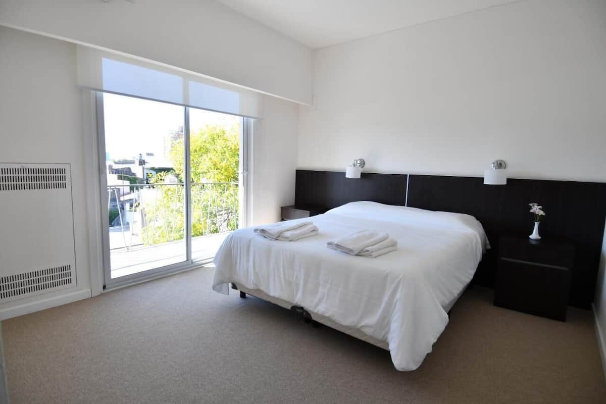 A hotel room with a large bed, a nightstand, a wall-mounted lamp, and a sliding glass door leading to a balcony.
