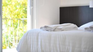 Una cama bien tendida con sábanas blancas y toallas dobladas, un cabecero oscuro y vistas a la vegetación a través de una ventana.