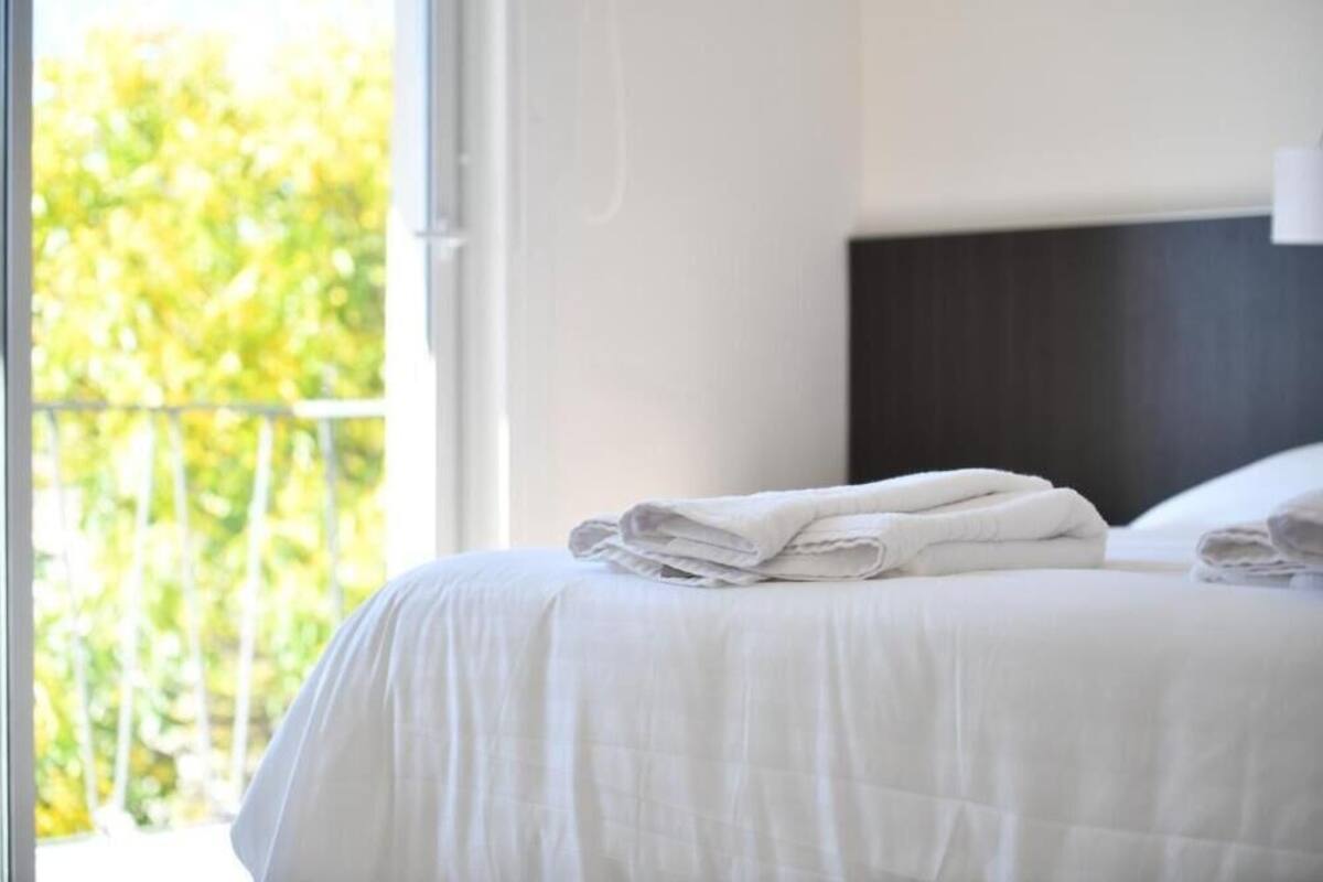 A neatly made bed with white linens and folded towels, a dark headboard, and a view of greenery through a window.