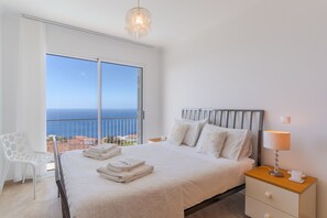 Ein Schlafzimmer mit einem großen Bett, einem Nachttisch, einem Sessel und Blick aufs Meer.
