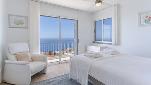Un dormitorio con cama, una silla, ventilador de techo y vistas al mar a través de amplios ventanales.