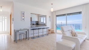 Una sala moderna con un sofá blanco, un comedor con taburetes de bar, una cocina con un ventanal grande y vistas al océano.