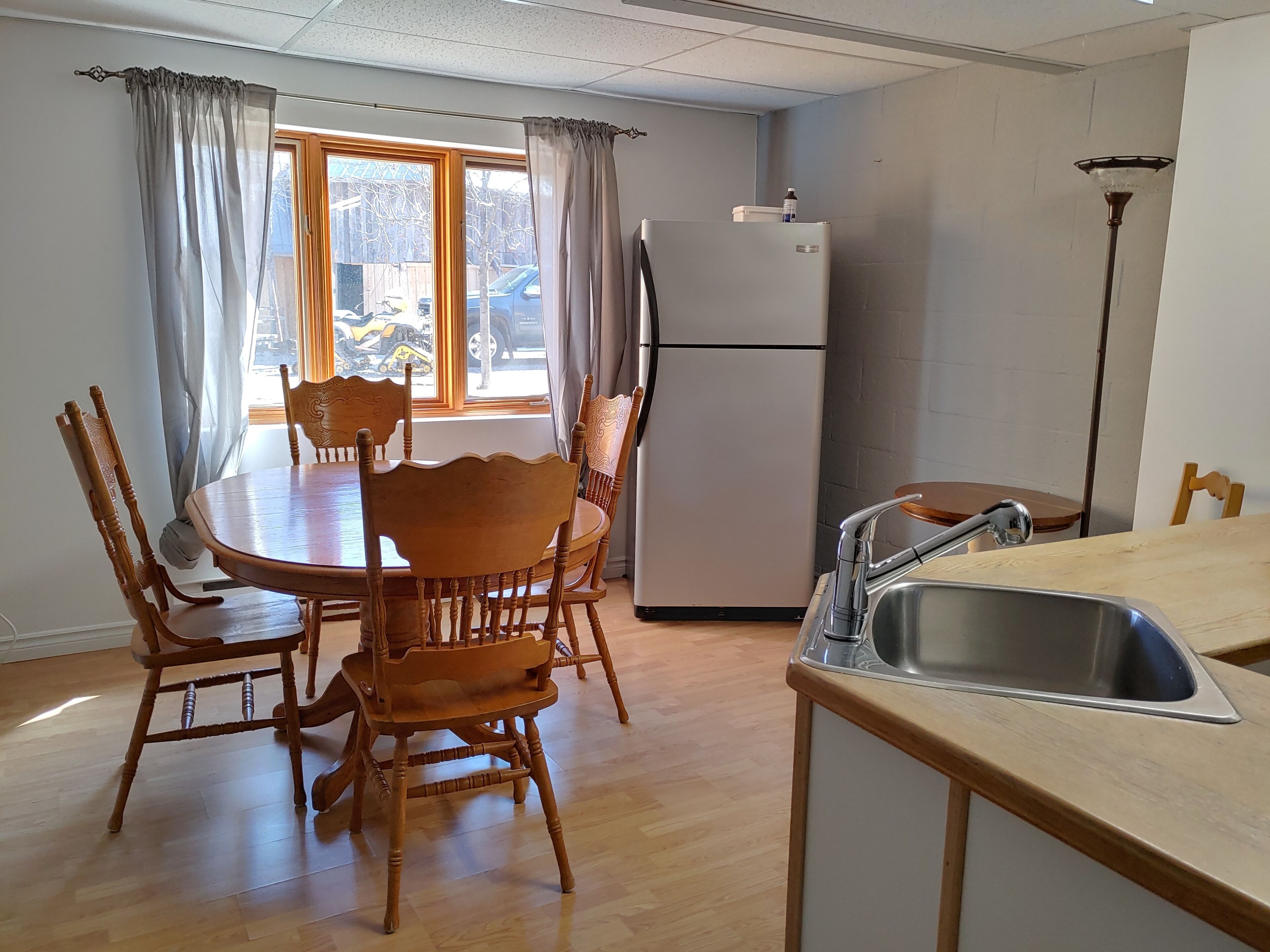 Un comedor con una mesa y sillas de madera, un refrigerador, un lavamanos y una ventana con cortinas.