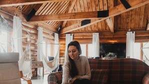 A cozy cabin interior with a wooden ceiling, a plaid sofa, a hammock, and a wooden table.