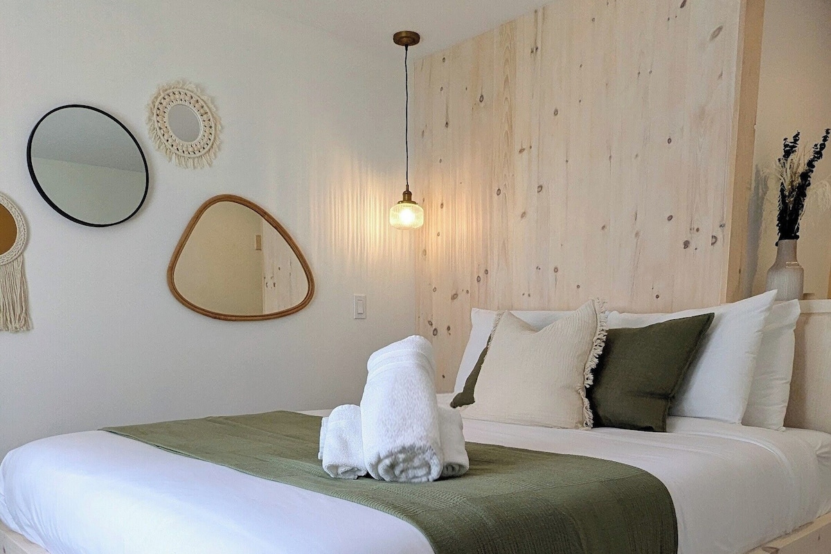 A wooden bed with white linens, a green throw blanket, and a towel. There are two wall-mounted mirrors, a woven light fixture, and a vase with dried plants.