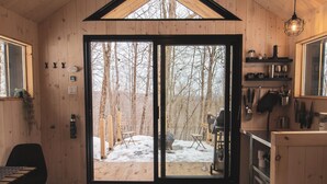A cozy cabin interior with a large bed, a dining area, and a view of a snowy forest through a glass door.