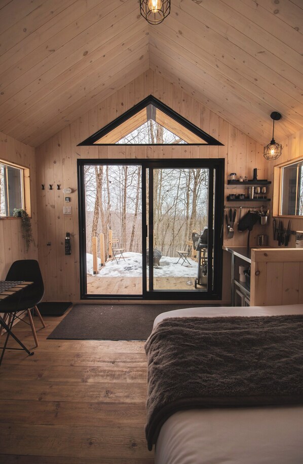 Interior acogedor de una cabaña con una cama grande, una zona de comedor y vistas a un bosque nevado a través de un ventanal.