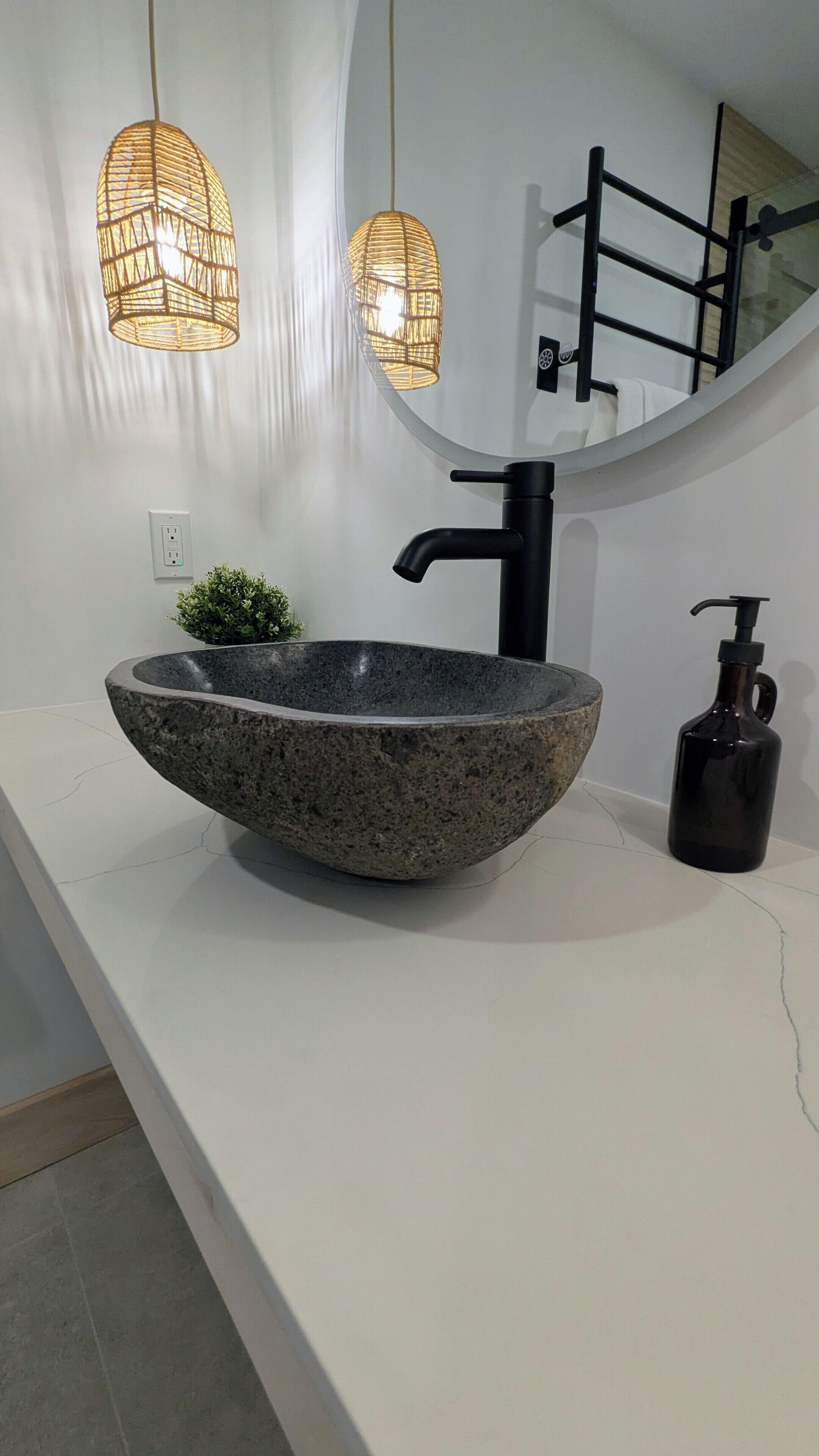 A modern bathroom with a large, oval-shaped stone sink, a black faucet, and a black soap dispenser. There are two woven light fixtures above the sink and a mirror with a black frame on the wall.