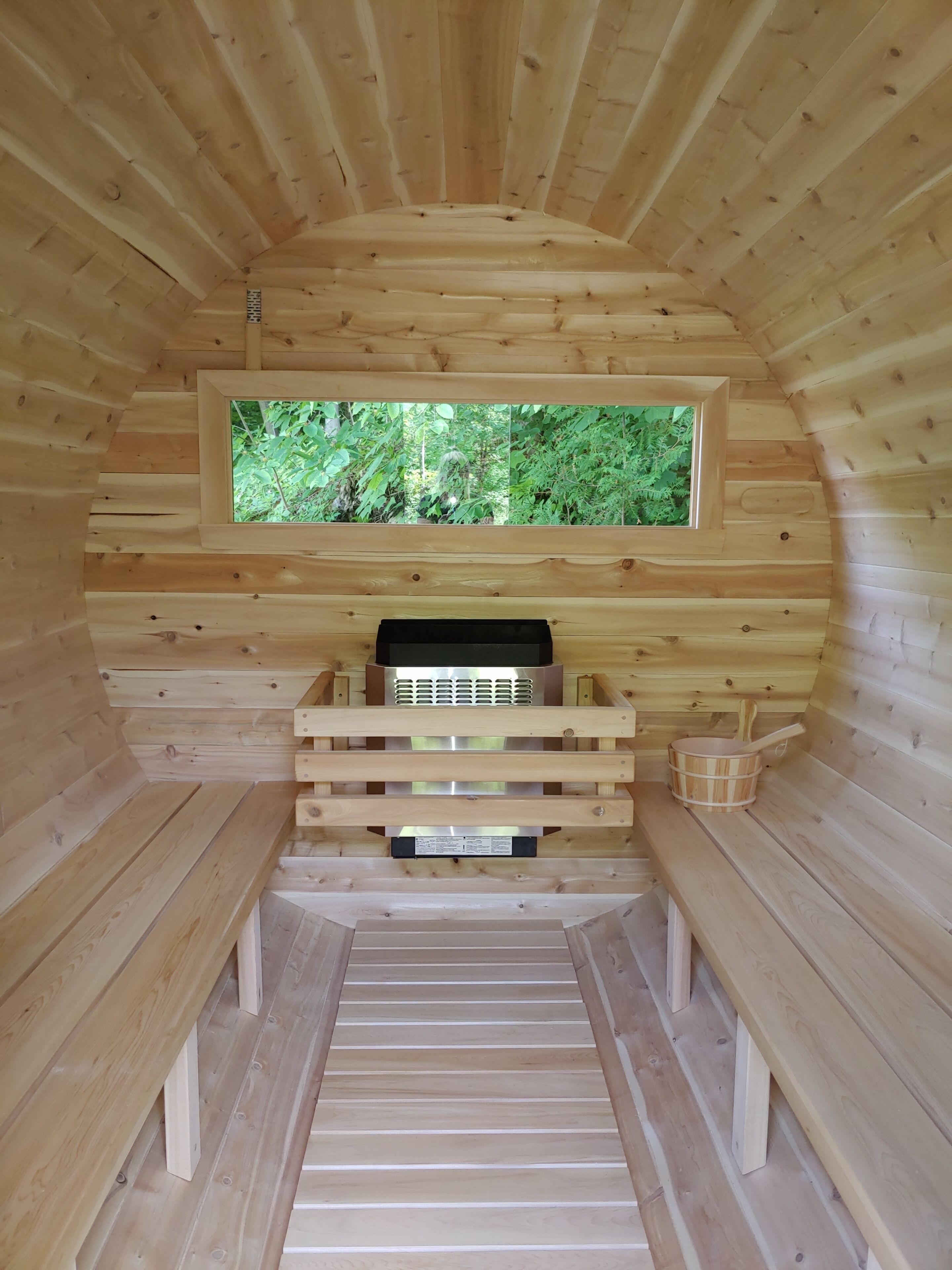 A sauna room with wooden walls, a bench, a sauna heater, and a window with a view of greenery.