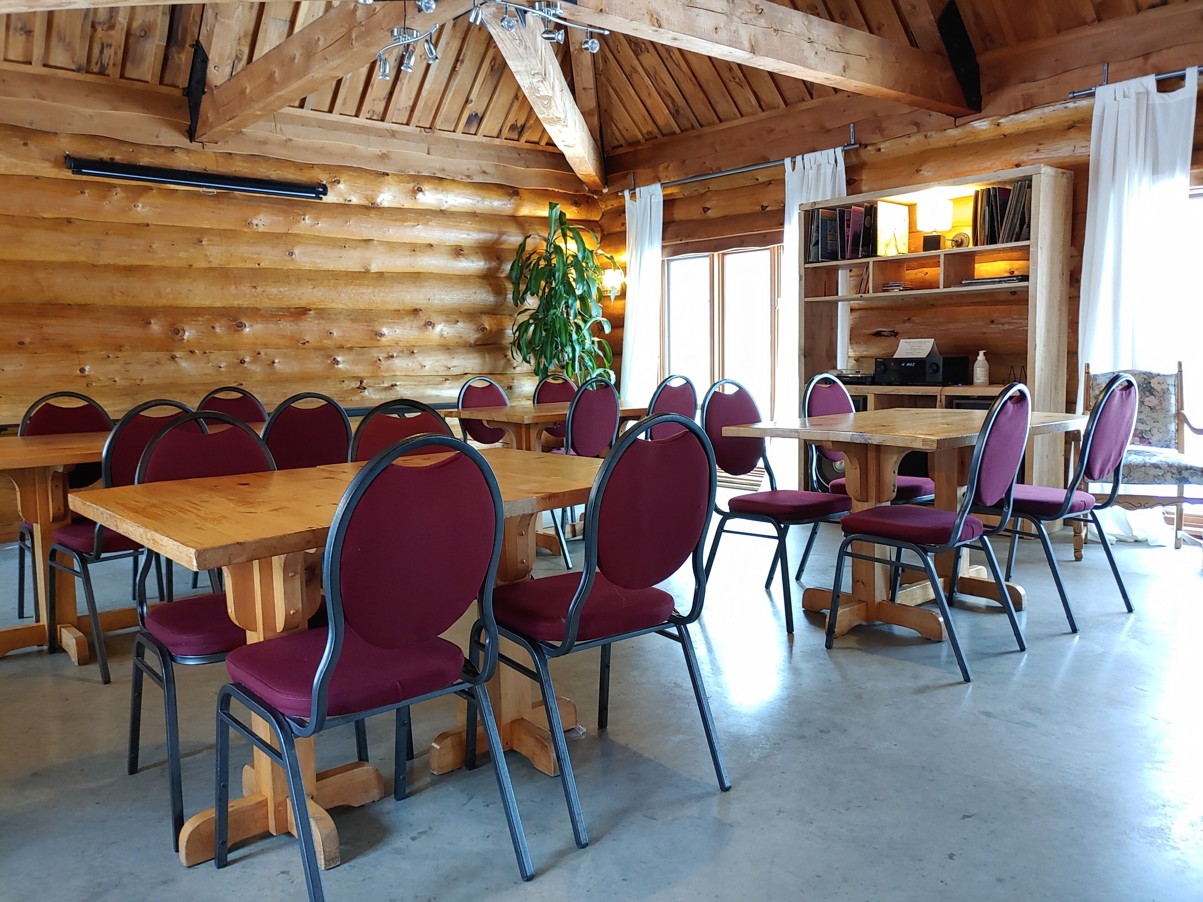 A wooden cabin interior with a long dining table, red cushioned chairs, and a bookshelf filled with books.