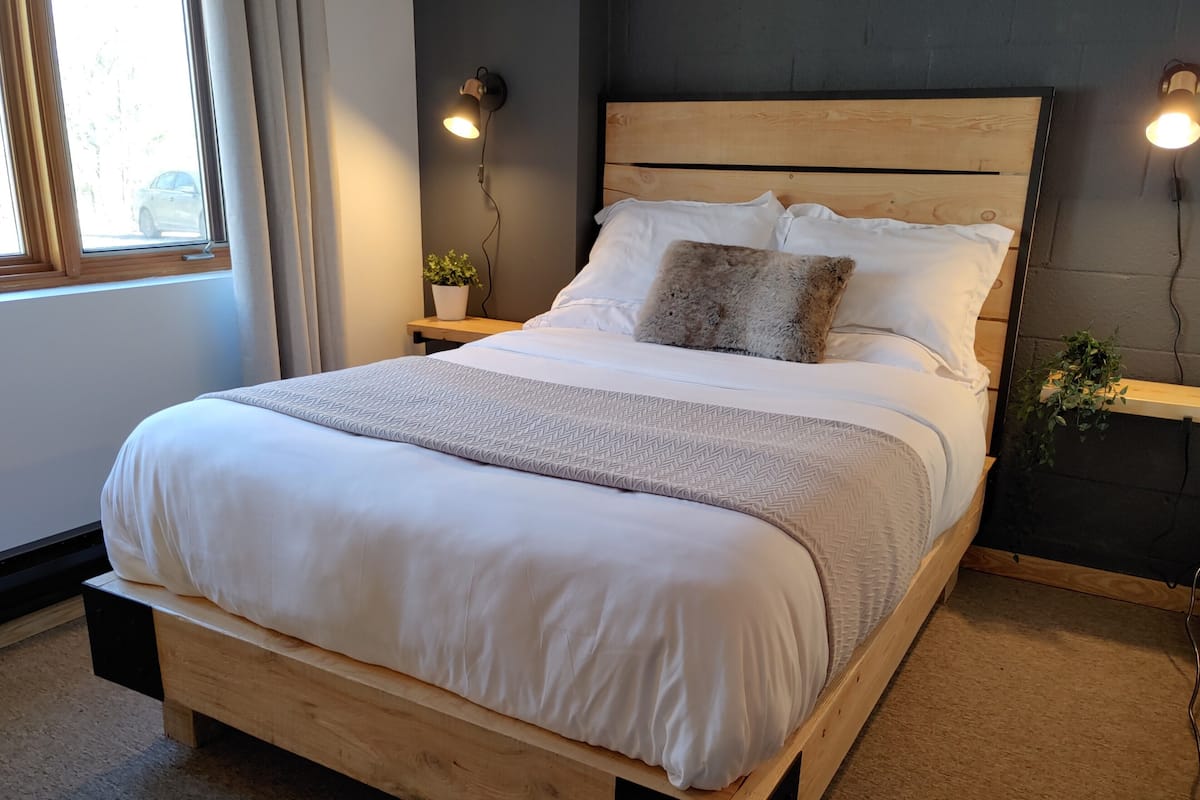 A neatly made bed with a wooden frame, white bedding, and a gray patterned throw. There are two bedside tables with lamps, a potted plant, and a view of the outside through a window.