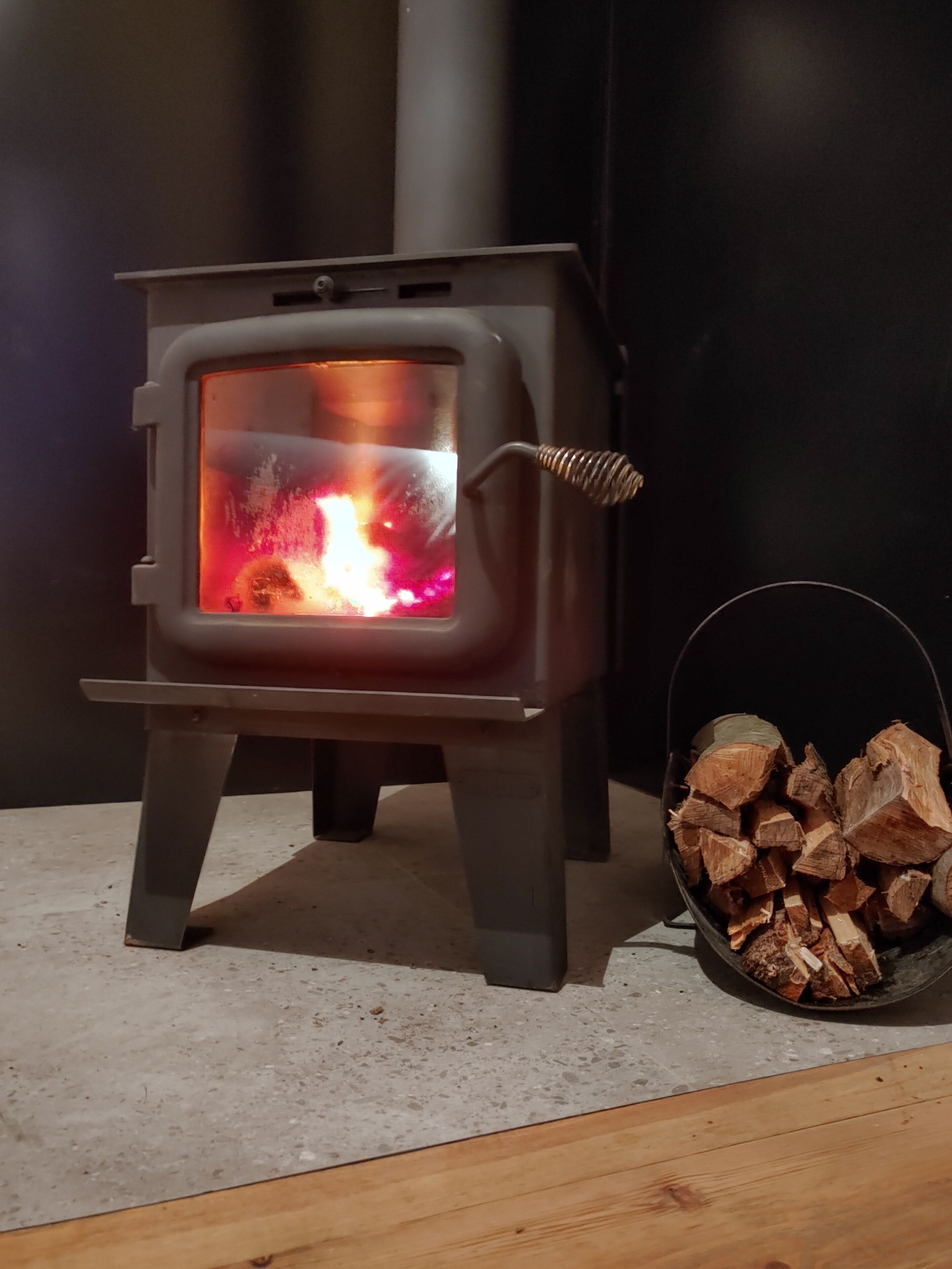 Ein Kaminofen mit sichtbarem Feuer, einem Metallständer und einem Holzkorb.
