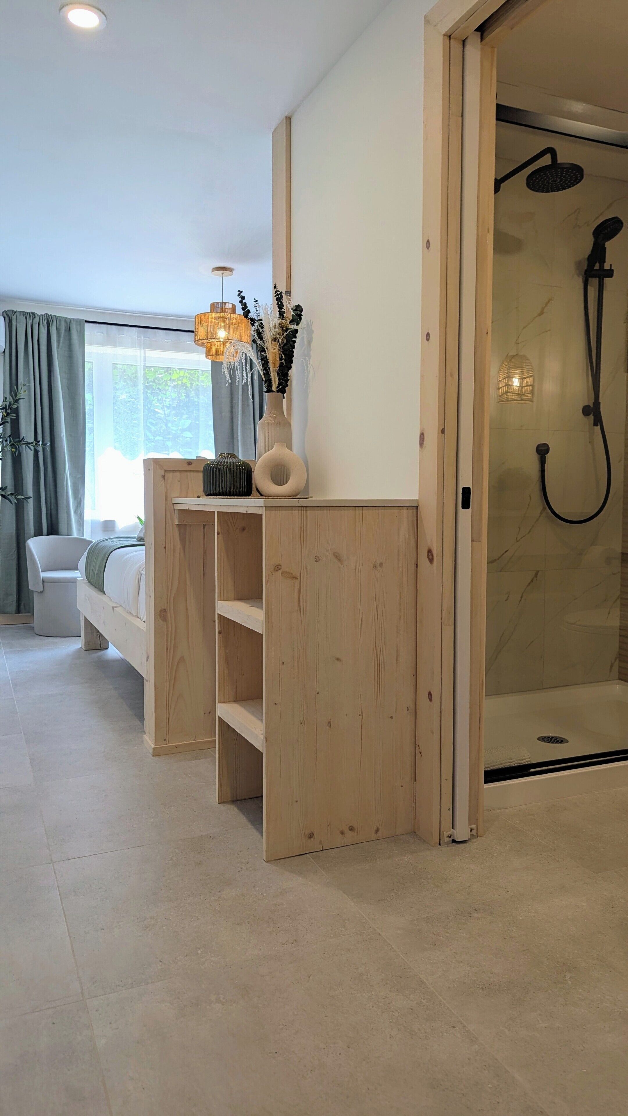 A modern bathroom with a glass shower enclosure, a wooden vanity with a vase of dried plants, and a bedside chair with a bed in the background.