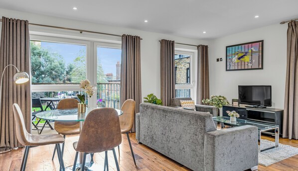 A modern living room with a glass dining table, comfortable seating, a flat-screen TV, and large windows with curtains.