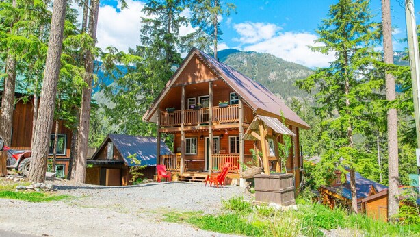 A wooden cabin with a balcony, surrounded by trees and a mountain backdrop.