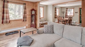 A cozy living room with a sofa, a wooden floor, a dining area with a table and chairs, and a large window with curtains.