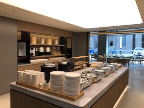 Un área de desayuno de hotel moderna con un sector de autoservicio, una máquina de café y vista a la calle.