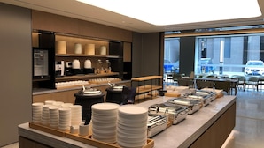 Un área de desayuno de hotel moderna con un sector de autoservicio, una máquina de café y vista a la calle.
