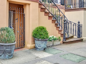 A building with a beige exterior, a metal gate, and a staircase with ornate black railings. There are potted plants in front of the building. - Romantic Garden Apartment on Chatham Square (Savannah)
