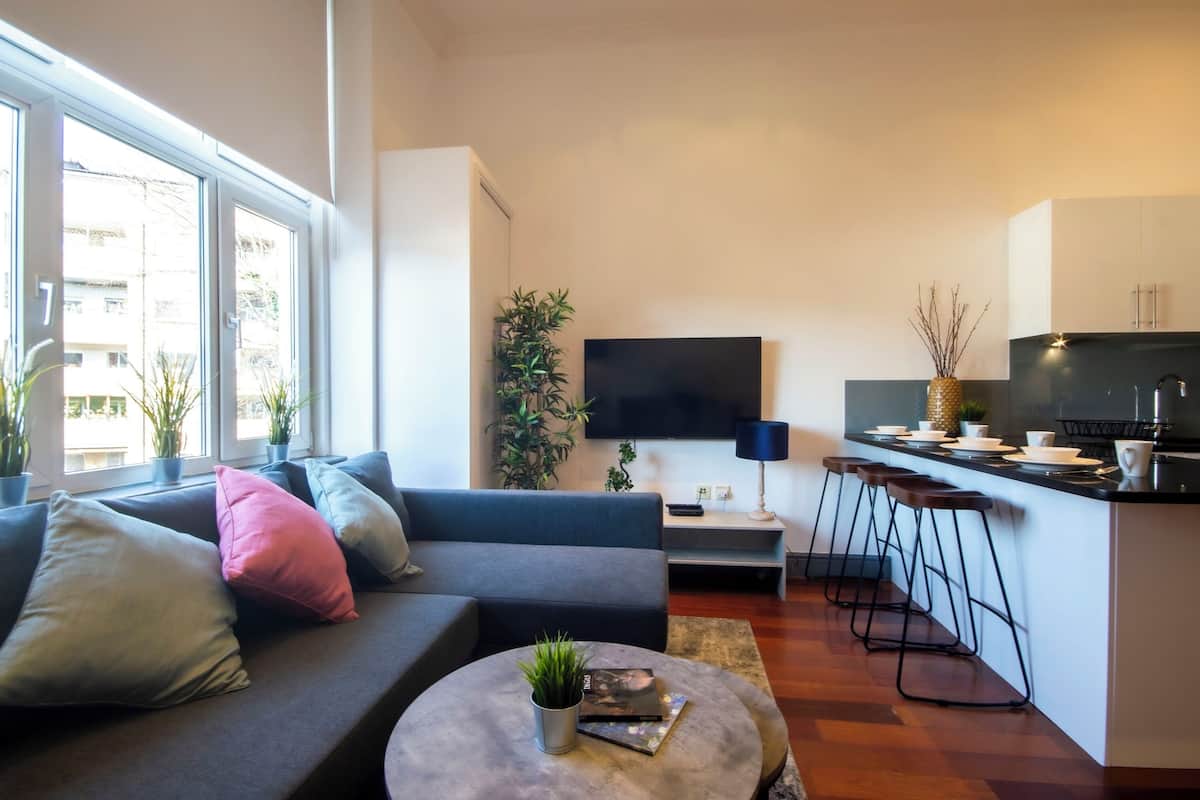 A modern living room with a sofa, a coffee table, and a TV. There is a kitchen area with a counter and stools.