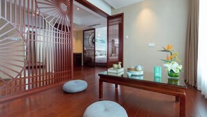 A room with a wooden floor, a glass table, two grey poufs, a decorative screen, and a vase with flowers. - Lanxin Hotel (Shaoxing)