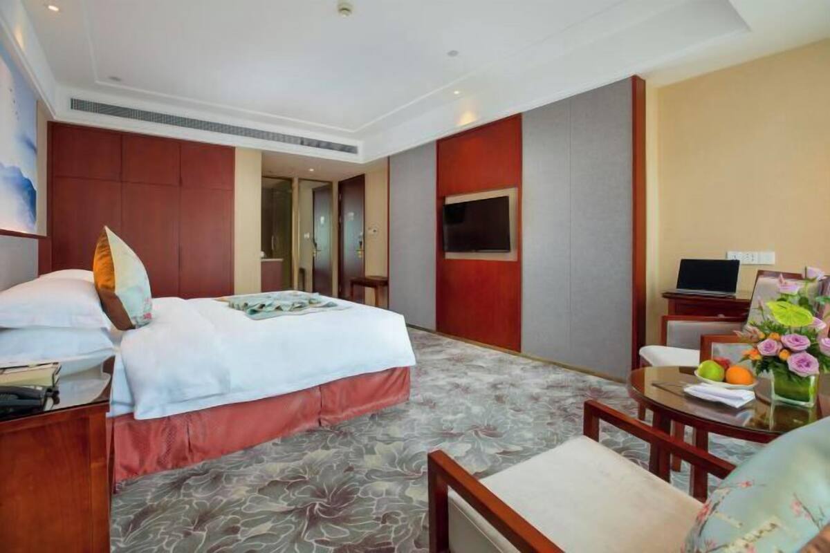 A hotel room with a bed, a desk, a chair, a TV, and a small table with a vase of flowers.