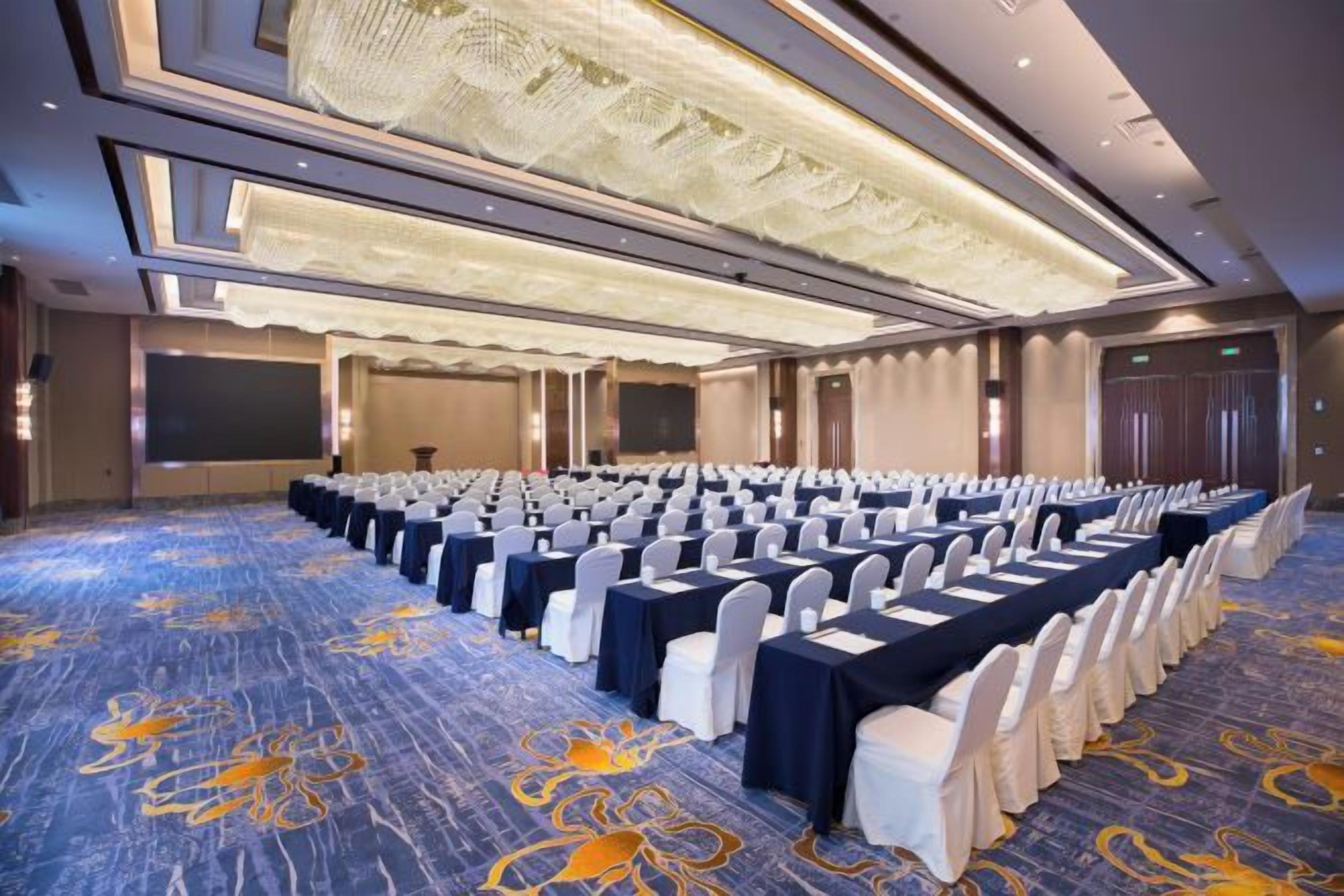 Un salón de conferencias con filas de mesas y sillas, piso alfombrado y una lámpara de araña grande.
