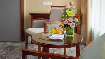 A hotel room with a round glass table, a chair, a vase with flowers, and a fruit bowl.