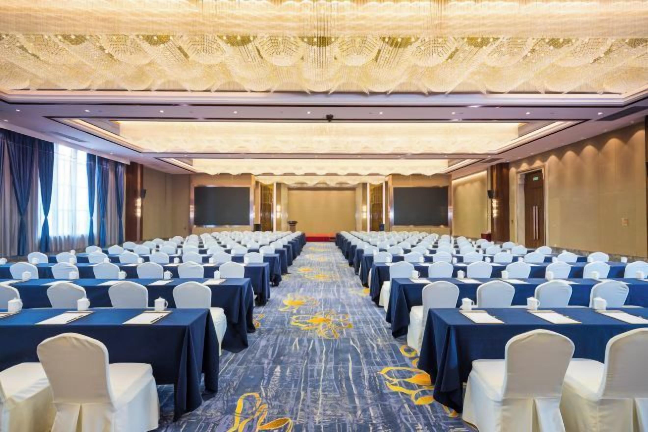 Un salón de conferencias con filas de mesas y sillas, piso alfombrado y techo con diseño.