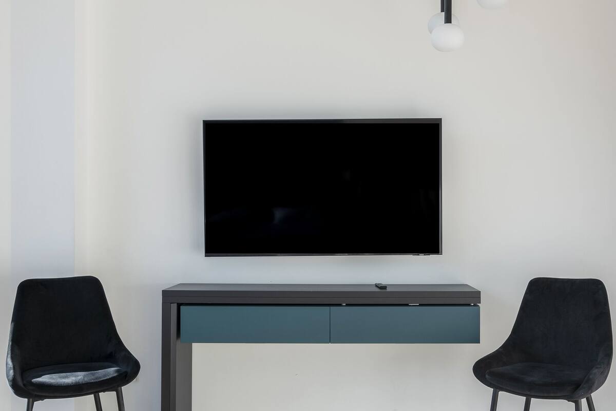 A modern room with a flat-screen TV mounted on a dark blue cabinet, two black chairs, and a simple pendant light fixture.