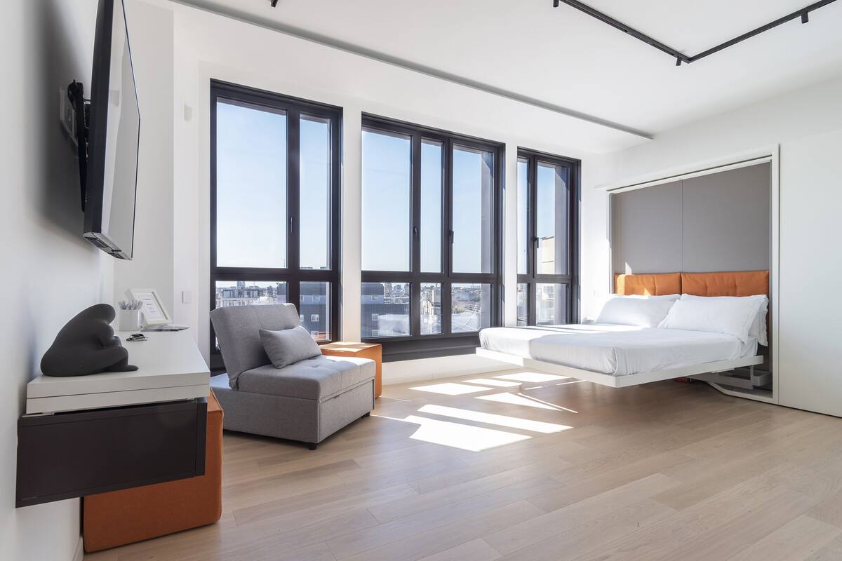 A modern hotel room with a bed, a sofa, a desk, and large windows offering a city view.