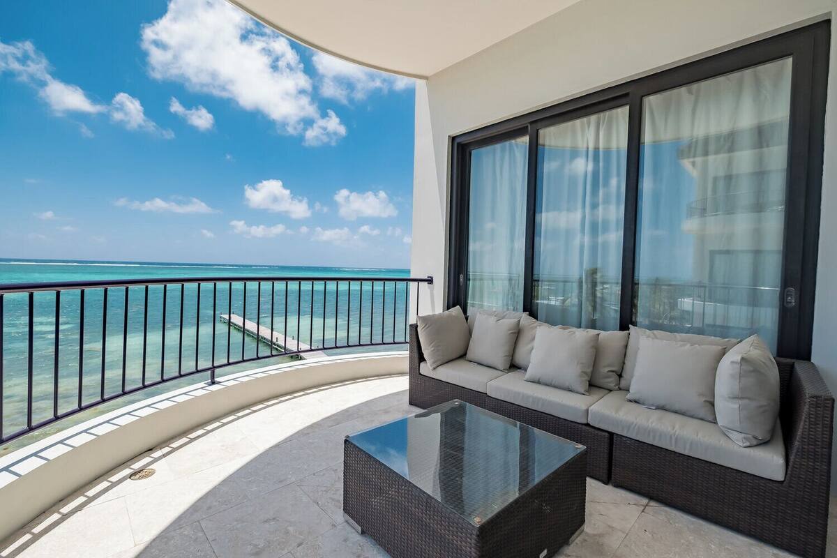 A balcony with a sofa and a glass table, overlooking a turquoise sea and a clear sky.