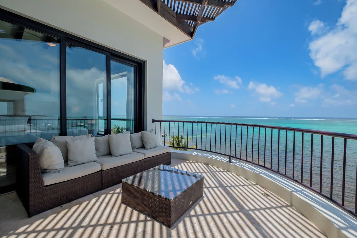 A balcony with a sofa and a glass coffee table, overlooking a turquoise sea.