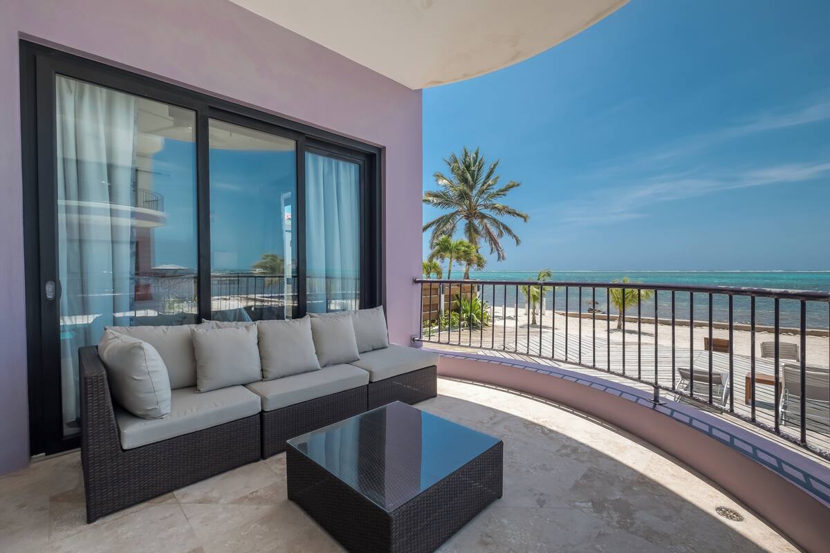 A balcony with a sofa and a glass-top coffee table, overlooking a beach with palm trees and the ocean.