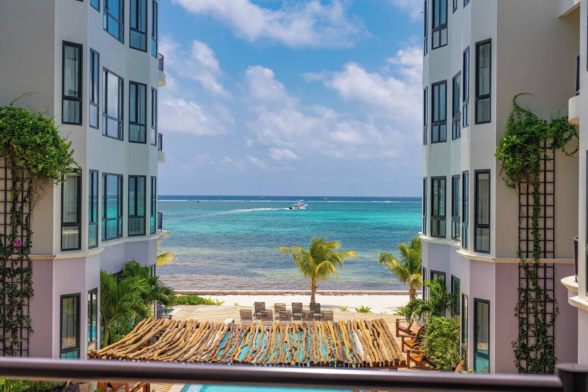 A view from a balcony overlooking a beach with a pool, lounge chairs, and a palm tree.