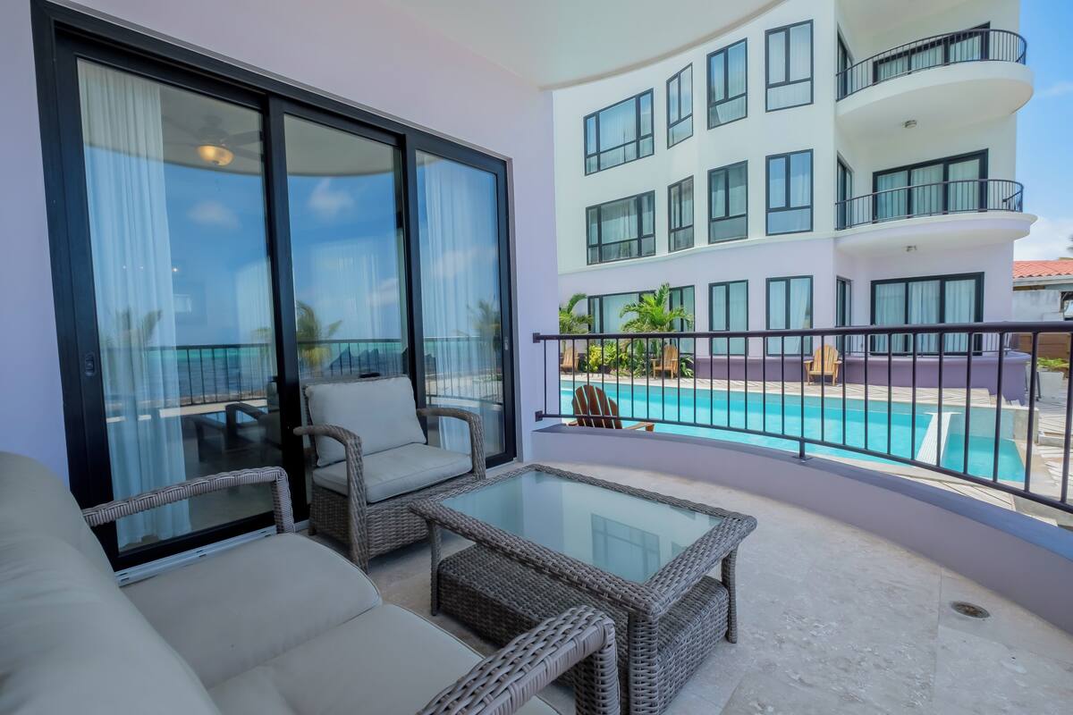 A balcony with wicker furniture, a glass table, and a view of a swimming pool and building.