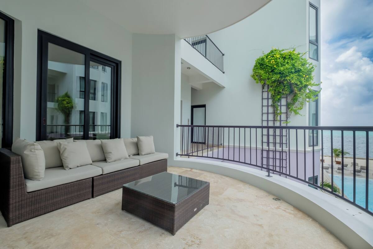 A modern living room with a sofa and a glass coffee table, a balcony with a railing, and a view of the sea.