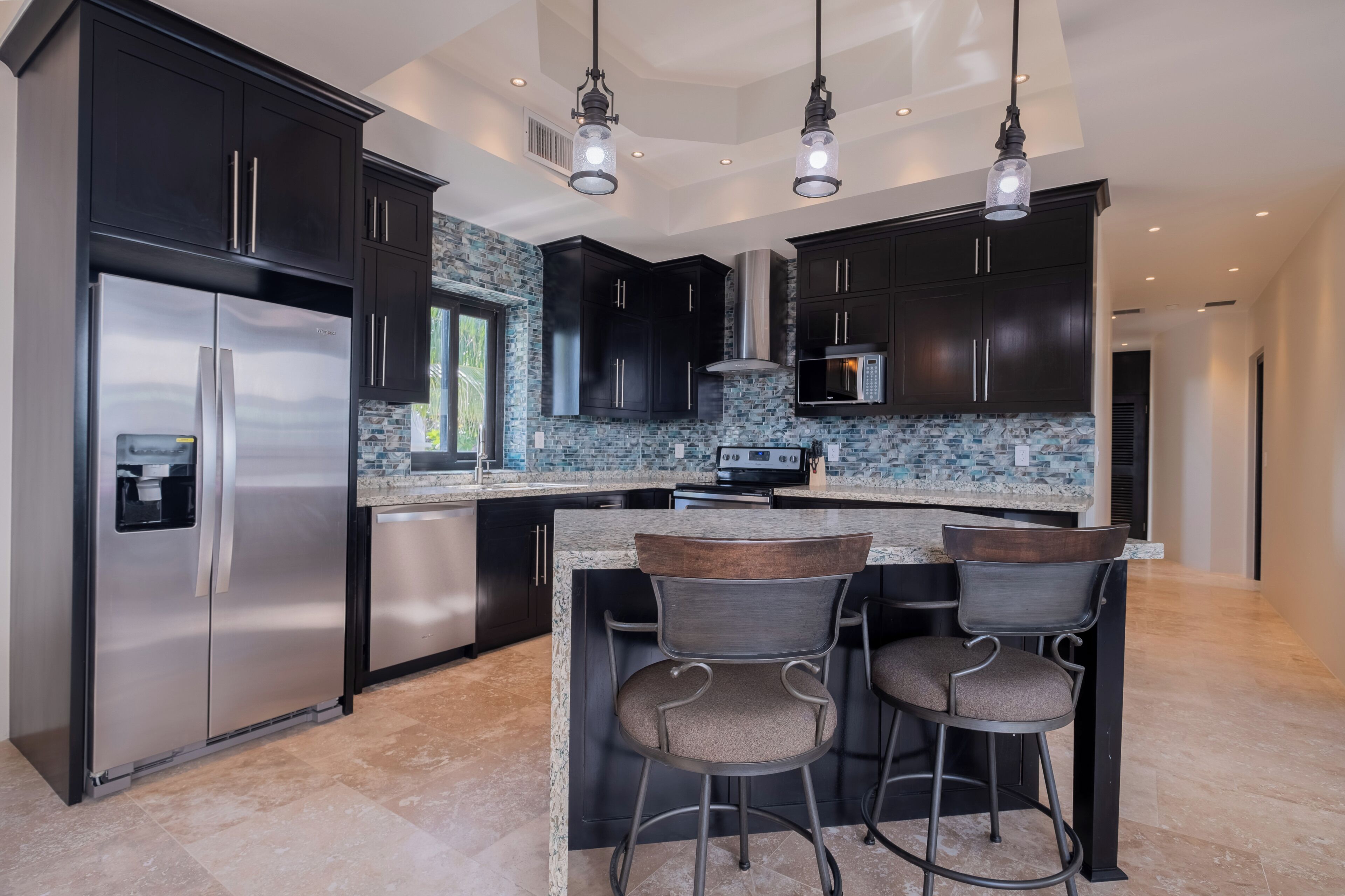 A modern kitchen with dark cabinetry, a stainless steel refrigerator, a granite countertop, and two bar stools.