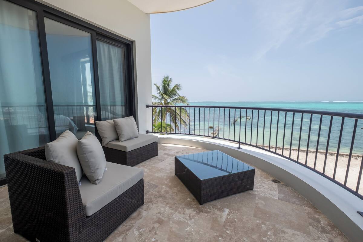A balcony with a view of the ocean, featuring wicker furniture and a glass table.