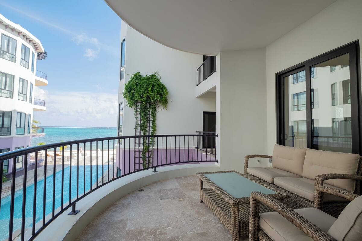 A balcony with a wicker sofa and a small table, overlooking a pool and a building with a balcony.