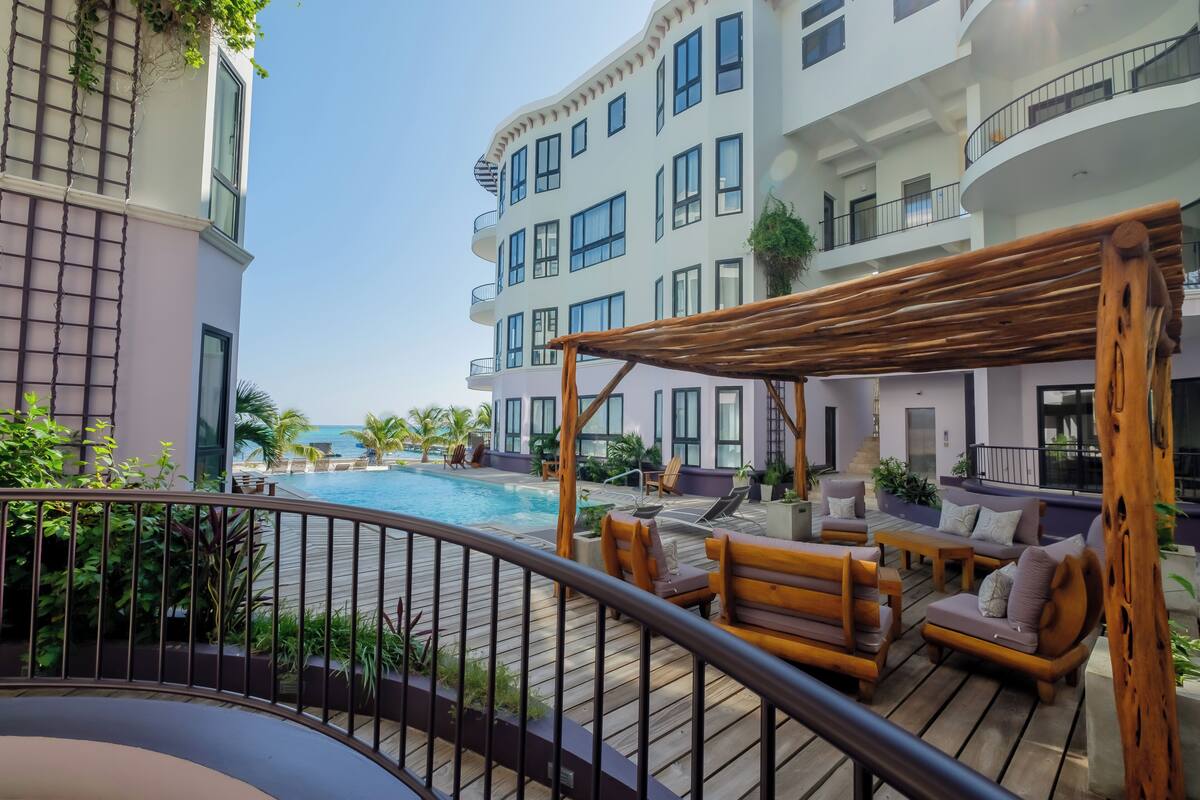 A poolside area with wooden deck furniture, a pergola, and a view of a multi-story building with balconies.