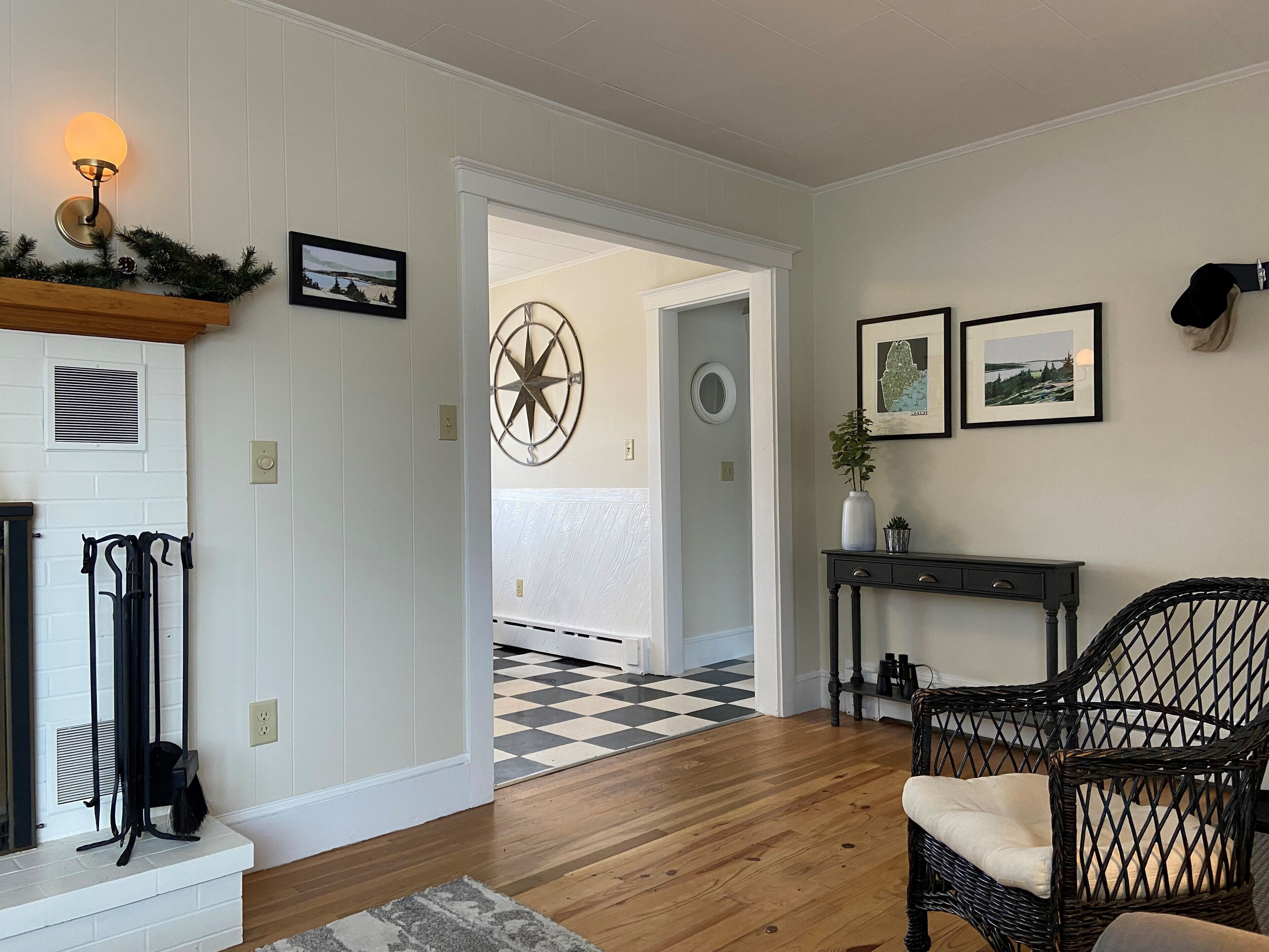 Un salon avec un sol à carreaux, une cheminée, une étoile décorative fixée au mur et une console sur laquelle sont posés un vase et des cadres photo.