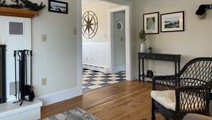Un salon avec un sol à carreaux, une cheminée, une étoile décorative fixée au mur et une console sur laquelle sont posés un vase et des cadres photo.