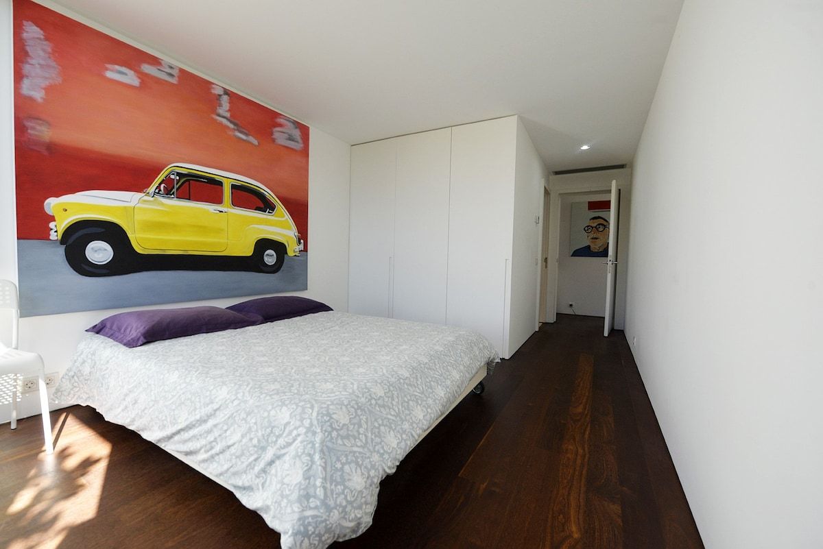 Een slaapkamer met een bed, een aan de muur hangend schilderij van een auto en een deur naar een andere kamer.