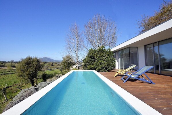 Una piscina infinita con cubierta de madera, dos sillas de descanso y vistas a un paisaje montañoso.