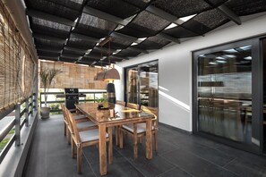 A modern outdoor dining area with a wooden table and chairs, a hanging pendant light, and a view of the city through large windows.