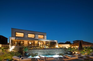 A modern two-story house with a swimming pool, outdoor seating, and a stone wall.