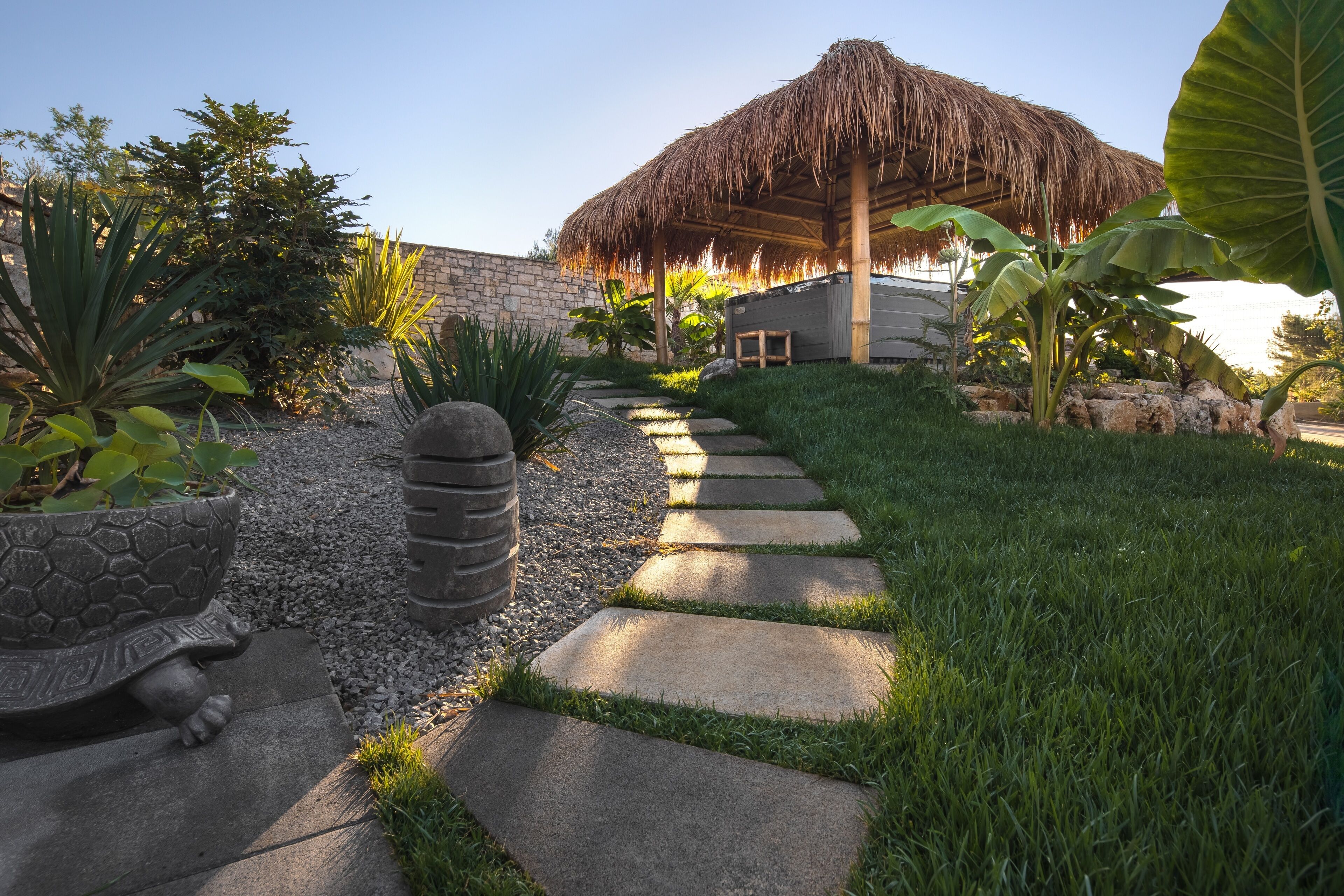 A garden pathway leading to a thatched-roof structure, surrounded by various plants and a stone wall.